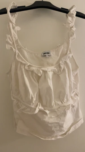 Vit volang linnetopp från Bubbleroom - Supersöt vit linnetopp från Bubbleroom i storlek L. Toppen har breda axelband med volanger och flera volangdetaljer framtill och runt midjan. Den är ärmlös och har en luftig, loose passform som ger en chill vibe. Perfekt för varma dagar och sommarhäng.