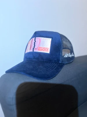 Blå meshkeps med 500-lapp patch - Snygg marinblå keps med mesh på sidorna och bak, samt en unik patch framtill med motiv av en svensk 500-kronorssedel. Kepsen har böjd skärm och broderad vit text på sidan. Perfekt för dig som gillar statement-plagg och streetstyle.