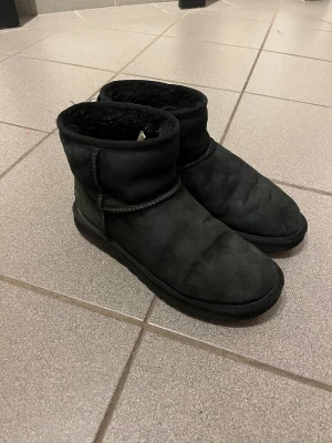 Svarta UGG boots i mocka - Mysiga svarta UGG boots med rund tå och platt sula. Utsidan är i mjuk mocka och insidan är fodrad med päls för extra värme. Klassisk UGG-logga baktill. Skorna är använda och fluffet på insidan har ”plattats till” något, annars bra skick.