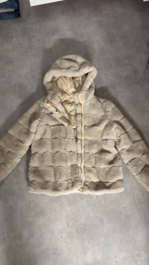Beige teddyjacka med huva - Mysig teddyjacka i beige med huva och quiltad insida. Jackan har dragkedja framtill och är långärmad. Perfekt för dig som gillar fluffiga och sköna jackor med chill vibe. Passar dig som vill ha en varm och snygg look under kalla dagar.