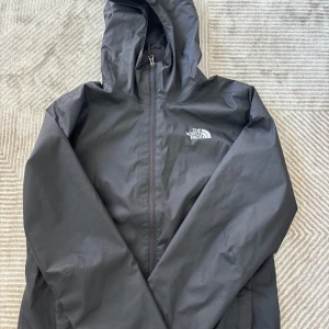 Svart vindjacka/regnjacka från The North Face - Svart vindjacka/regnjacka från The North Face med huva och dragkedja framtill. Jackan har en clean look med logga på bröstet och praktiska fickor. Perfekt för blåsiga dagar och enkel att matcha med allt. Snygg och funktionell för dig som gillar streetwear. Pris kan diskuteras!!