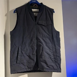 Mörkblå dunväst från Hilfiger Denim - Snygg mörkblå dunväst från Hilfiger Denim med dragkedja framtill, bröstficka med lock och två sidofickor. Västen har en enkel, quiltad design och står för en clean och sportig look. Perfekt för lager-på-lager-stil under kyliga dagar.