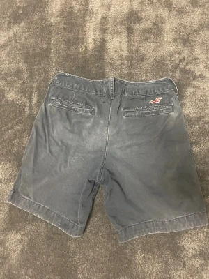 Hollister shorts - Snygga mörkblåa shorts från Hollister i storlek  W28 med klassisk passform och midwaist. De har två fickor bak, en med broderad Hollister-logga, och två fickor fram. Materialet är bomull och de stängs med knappar. Skicka ett dm vid minsta beundran. Priset är ej hugget i sten.