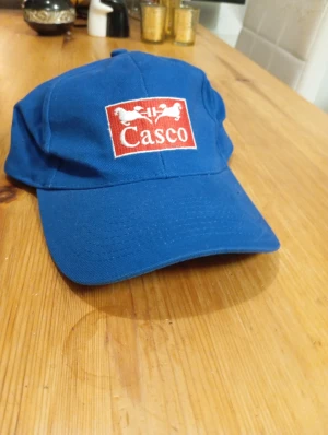 Blå Casco keps med broderad logga - Snygg blå keps från Casco med broderad röd och vit logga framtill. Klassisk form med böjd skärm och justerbar passform. Perfekt för dig som gillar sportig stil och vill sticka ut med en unik accessoar.