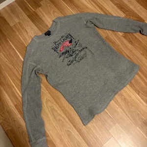 Grå långärmad tröja Polo Ralph Lauren - Säljer en grå långärmad tröja från Polo Ralph Lauren med tryck framtill och amerikansk flagga. Tröjan är ribbad och har rund hals. Perfekt för dig som gillar klassisk streetstyle och vill ha något bekvämt och snyggt. Kan gå ner i pris under snabb affär!