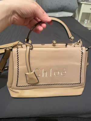 Beige axelväska och plånbok från Chloé - Snygg beige axelväska från Chloé med bred axelrem med guldfärgade öljetter och matchande plånbok. Väskan har ett stilrent broderat Chloé-logo framtill, svarta kanter och gulddetaljer. Både väska och plånbok är i mjukt skinn med minimalistisk design. Köpt sacond hand men i fint skick utan defekt