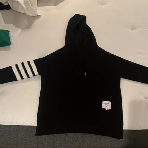 Svart hoodie med vita ränder från Thom Browne - Svart hoodie från Thom Browne med huva och snörning. Ena ärmen har fyra vita, breda ränder som ger en sportig vibe. Långärmad och med en vit etikett med märkets logga nedtill. Stickad i mjukt material, perfekt för chill dagar.