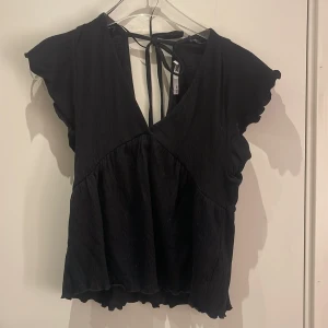 Svart blus med knyt från 157 - Svart blus från 157 med v-ringning och snygg knytning i ryggen. Modellen har korta, vågiga ärmar och volangkant nedtill. Materialet är lätt och har en skön struktur som ger en cool look. Perfekt för dig som gillar stilrena och trendiga plagg.