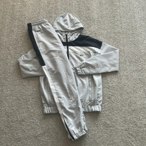 Lacoste grå och svart tracksuit - Säljer en stilren tracksuit från Lacoste i ljusgrått med svarta detaljer. Setet består av en jacka med huva, dragkedja och meshfoder samt matchande byxor med resår i midja och benslut. Ikoniska Lacoste-loggan syns på både jacka och byxa.