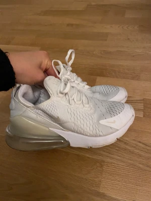 Nike Air Max 270 vita sneakers - Säljer ett par fräscha Nike Air Max 270 i helvitt med stora luftbubblor i hälen. Skorna har mesh-överdel, snörning och diskreta swoosh-loggor. Perfekta för dig som gillar sportig och clean stil. Supersköna och lätta på foten.