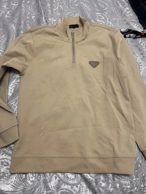 Beige half zip tröja från Prada - Snygg beige långärmad tröja från Prada med half zip framtill och diskret logga på bröstet. Tröjan har en clean look med ribbade muddar och är gjord i mjuk bomull. Perfekt för dig som gillar stilrena och exklusiva plagg.