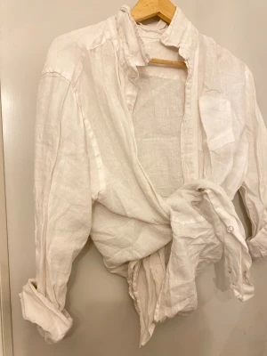 Vit linneskjorta med knyt framtill - Snygg vit skjorta i linne med knyt framtill och uppvikta ärmar. Skjortan har en klassisk krage, knappar och en bröstficka. Perfekt för dig som gillar en chill och avslappnad stil med lite edge.