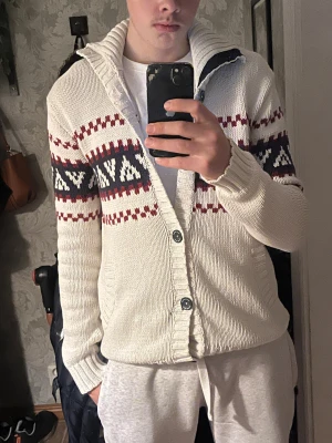 Vit stickad kofta med mönster - Säljer en vit stickad kofta med snyggt mönster i rött och mörkblått över bröst och armar. Koftan har knappar framtill, ribbade muddar och en mysig krage. Perfekt för lager-på-lager och chill dagar.