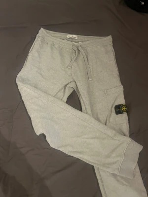 Grå mjukisbyxor från Stone Island - Snygga grå mjukisbyxor från Stone Island med klassisk logopatch på benet. Byxorna har snörning i midjan, ribbade muddar och en praktisk ficka på sidan. Perfekta för chill dagar och har en clean, modern look. Nya kostar 3200kr men kom gärna med förslag på priset 