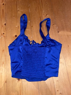 Blå satin korsett-topp med snörning - Snygg blå korsett-topp i satin med breda axelband och smockad framsida. Toppen har snörningsdetalj vid bysten och är croppad i modellen. Perfekt för fester , använder inte ofta mera då de inte e min style ❤️❤️