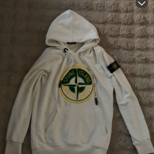  Stone Island hoddie  - Vit hoodie från Stone Island med stort broderat logomärke i grönt och gult på bröstet samt klassisk patch på ärmen. Tröjan har huva med snörning och känguruficka framtill. Tillverkad i 100% bomull för en skön och avslappnad känsla.