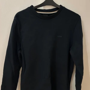 Mörkblå sweatshirt från BOSS - Mörkblå sweatshirt från BOSS med diskret logga på bröstet. Tröjan har rund hals och långa ärmar, samt en enkel och stilren design. Materialet är mjukt och känns skönt mot huden, perfekt för dig som gillar klassisk streetwear.