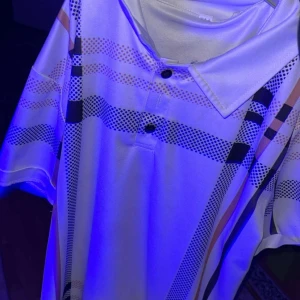 Vit rutig henley skjorta med knappar - Snygg vit henley skjorta med rutigt mönster i svart och beige. Skjortan har korta ärmar, klassisk krage och två knappar framtill. Materialet känns slätt och har en lätt glans, vilket ger en fräsch vibe. Perfekt för dig som gillar stilrena detaljer och mönster.