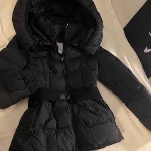 Svart Moncler dunjacka med bälte - Vintage vinterjacka från Moncler. Köptes tidigare  på Vinted. Väldigt snygg med fina detaljer kring midjan och hals. Även stor och härlig luva.   I använt skick men inga hål Storlek s på taggen.