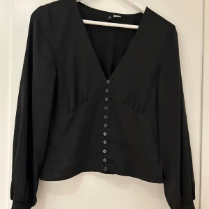 Svart topp strl 38 - Snygg svart blus med djup v-ringning med knappar längs framsidan. De. Är sparsamt använd och sitter fint. Den är i storlek 38