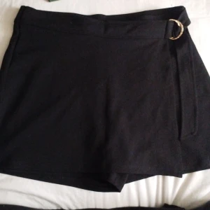 Svarta shorts med bälte från H&M - Snygga svarta shorts från H&M i storlek M. De har hög midja och ett brett bälte med en guldig ringdetalj framtill. Materialet är mjukt och känns som polyester. Passformen är normal och de är stilrena utan mönster, perfekta för att lyfta din outfit. Det ser ut som en kjol. 