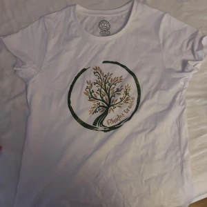 Vit t-shirt med trädtryck Rhodes Grove - Vit t-shirt från Rhodes Grove med ett stort tryck av ett träd i grönt och guld på bröstet. Rund halsringning och korta ärmar. Skön och mjuk i bomull, perfekt till jeans eller shorts. Vill helst ha den här tröjan bort fort som möjligt 