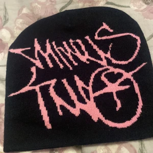 Svart mössa med rosa graffiti-tryck - Cool svart mössa med stort rosa graffiti-inspirerat tryck framtill. Mössan är stickad och har en klassisk, enkel form utan tofs eller andra detaljer. Perfekt för dig som vill sticka ut med streetstyle-vibe.