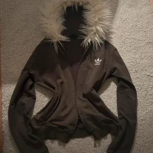 Adidas hoodiejacka med fuskpäls - Oliv färgad hoodiejacka från Adidas med vit fuskpäls runt huvan och klassiska vita ränder längs ärmarna. Jackan har dragkedja framtill, ribbade muddar och Adidas-logga på bröstet. Perfekt för dig som gillar alternativa kläder