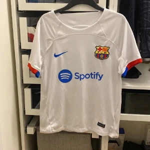 Vit FC Barcelona Nike fotbollströja S - Säljer en vit FC Barcelona fotbollströja från Nike med Dri-FIT-material. Tröjan har klubbmärke, blå Nike-logga och Spotify-tryck på bröstet samt UNHCR-tryck på ryggen. Ärmarna har röda och blå detaljer. Perfekt för dig som älskar Barca och snygg sportstil.