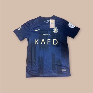 Marinblå Nike fotbollströja Al Nassr - Snygg marinblå fotbollströja från Nike för Al Nassr med guldiga detaljer och tryck. Tröjan har korta ärmar, rund hals och flera sponsorloggor på bröstet och ärmarna. Tillverkad i lätt och ventilerande polyester, perfekt för match eller träning.