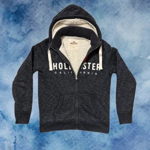 Fodrad Hollister Kofta - Mysig grå hoodie från Hollister med vit och röd logga på bröstet. Tröjan har dragkedja, huva och svart teddyfoder på insidan. Perfekt för kyliga dagar och har två fickor framtill. Längd ≈64 cm, Bredd Amrhåla - Armhåla ≈ 52 cm. Hör av vid frågor, priset ej hugget i sten!!