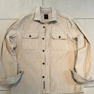 Beige overshirt från Lost Minds L - Snygg beige overshirt från Lost Minds i storlek L. Tillverkad i mjuk manchester med diskreta ränder, två stora bröstfickor med knappar och klassisk krage. Perfekt att slänga över en t-shirt för en chill look. Långa ärmar och svarta knappar framtill.