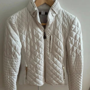 Creamvit quiltad jacka från Zara - Creamvit quiltad bombe jacka från Zara med snygga guldfärgade dragkedjor och ribbade sidor. Jackan har ståkrage, lång ärm och flera fickor med dragkedja. Perfekt för dig som gillar stilen och sportig look