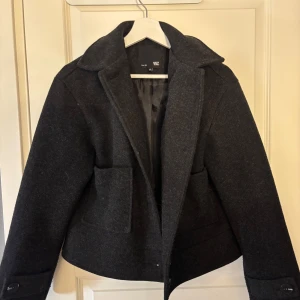 Mörkgrå kappa från 157 - Snygg mörkgrå kappa från 157 med klassisk krage och stora fickor framtill. Jackan är tillverkad i ull och har en stilren, rak passform som passar perfekt till höstens och vinterns outfits. Knappar vid ärmslut och fodrad insida för extra komfort.