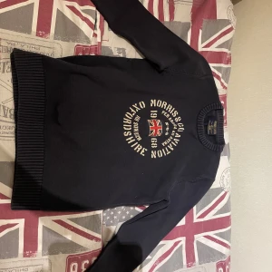 Mörkblå stickad tröja Morris - Snygg mörkblå stickad tröja från Morris med rund hals och ribbade muddar. Framsidan har ett stort tryck med brittisk flagga och text i vitt. 