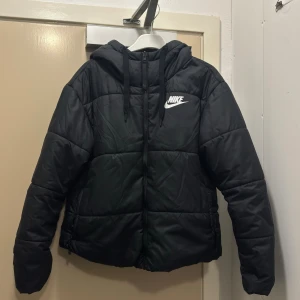 Svart pufferjacka från Nike - Svart pufferjacka från Nike med huva och snörning. Jackan har en vit Nike-logga på brösten.