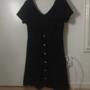 Svart kortärmad klänning från ONLY - Svart kortklänning från ONLY med v-ringning och knyt framtill. Klänningen har korta ärmar och dekorativa knappar längs hela framsidan. Enkel och stilren modell som passar perfekt till sommaren.