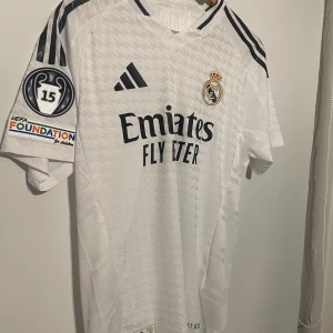 Real Madrid Mbappé 9 matchtröja slim fit  - Säljer en vit Real Madrid fotbollströja från Adidas med Mbappé och nummer 9 på ryggen. Tröjan har korta ärmar, svarta detaljer och klubbmärke på bröstet. Materialet är lätt och andas, perfekt för match eller träning. UEFA Foundation-märke på ärmen.