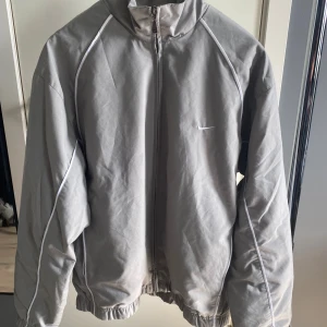 Grå vindjacka från Nike i storlek L - Säljer en stilren grå vindjacka från Nike i storlek L. Jackan har vita detaljer längs sömmarna, dragkedja framtill och elastiska muddar vid ärmslut och nederkant. Perfekt för dig som gillar sportig och clean stil.