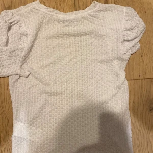 Vit blus med struktur från VILA - Säljer en vit blus från VILA i storlek XS. Toppen har korta puffärmar och ett snyggt strukturerat tyg som ger en cool och modern känsla. Rund halsringning och lätt transparent material. Perfekt för dig som gillar stilrena och trendiga plagg.