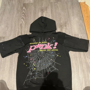 Svart hoodie från Spöder med tryck - Cool svart hoodie från Spöder med stort spindelnäts- och stjärntryck framtill, texten 'pråk!' i rosa och gult samt en gul stjärna på ryggen. Tröjan har huva och är gjord i 100% bomull. Perfekt för dig som gillar unika streetwear-plagg.
