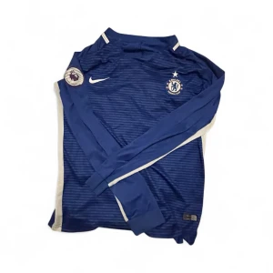 Chelsea långärmad fotbollströja Nike - Chelsea FC långärmad matchtröja från Nike i blått med tunna horisontella ränder och vita detaljer längs sidorna. Tröjan har klubbmärke och Premier League-patch på ärmen. Tillverkad i 100% polyester, perfekt för träning eller match.