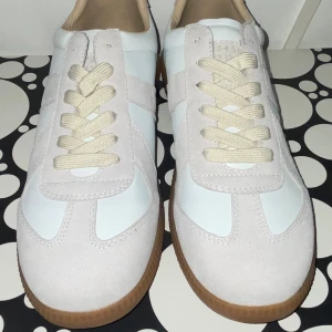 Mason margiela vita - Snygga sneakers i vitt med beige mockadetaljer och krämfärgade snören. Skorna har en klassisk design med rund tå och platt gummisula i brunt. Perfekta för dig som gillar stilrena och fräscha sneakers med retrovibbar.