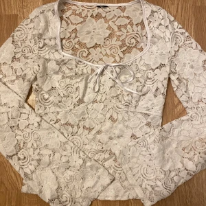 Vit spetsblus med knyt i halsen - Säljer en vit blus i spets med blommigt mönster och lång ärm. Toppen har en snygg fyrkantig ringning med knytband framtill och är genombruten i materialet. Perfekt för dig som gillar romantisk och feminin stil.