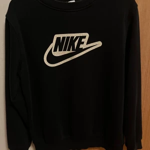 Svart Nike sweatshirt med logga - Svart sweatshirt från Nike med stor, vit logga på bröstet. Tröjan har rund hals och långa ärmar, perfekt för en chill och sportig stil. Materialet känns mjukt och skönt mot huden, och passformen är normal. En klassiker för dig som gillar streetwear.