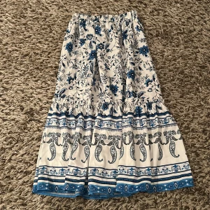 Vit och blå långkjol med blommönster - Säljer en super söt långkjol i vitt med blått mönster. Kjolen har volang och resår i midjan för skön passform. Materialet känns lätt och luftigt, perfekt för varma dagar. Mönstret är detaljerat med bla blommor.💗