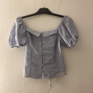 Blårandig blus med puffärmar - Säljer en blårandig blus med puffiga kortärmar. Toppen har knappar framtill och snörning i ryggen för extra snygg passform. Tillverkad i mjuk bomull. Söt krage och figurnära siluett.
