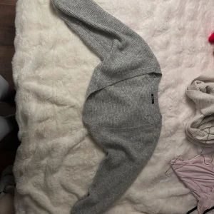 Grå ribbad stickad kofta från Gina Tricot - Säljer en grå stickad kofta från Gina Tricot med ribbad struktur. Koftan har långärmade ärmar och en enkel, klassisk design utan knappar eller dragkedja. Perfekt att slänga över en t-shirt för extra värme under kyliga dagar.