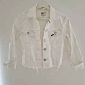 Vit jeansjacka från Lee, XS - Snygg vit jeansjacka från Lee i storlek XS. Jackan har klassisk krage, silverfärgade knappar, två bröstfickor med lock och knappar samt rå, fransig nederkant för en cool vibe. Kort modell och tillverkad i bomull. Perfekt att styla med dina favoritjeans. Tyget är något gulnat i nacken, men annars i fint skick.