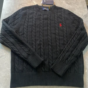 Svart kabelstickad tröja Polo Ralph Lauren - Svart kabelstickad tröja från Polo Ralph Lauren med klassisk rund hals och röd broderad logga på bröstet. Tröjan har ribbade muddar vid ärmslut och nederkant, samt långärmad design. Perfekt för dig som gillar stilren och tidlös look. Priset är inte hugget i sten så kom gärna med pris förslag.